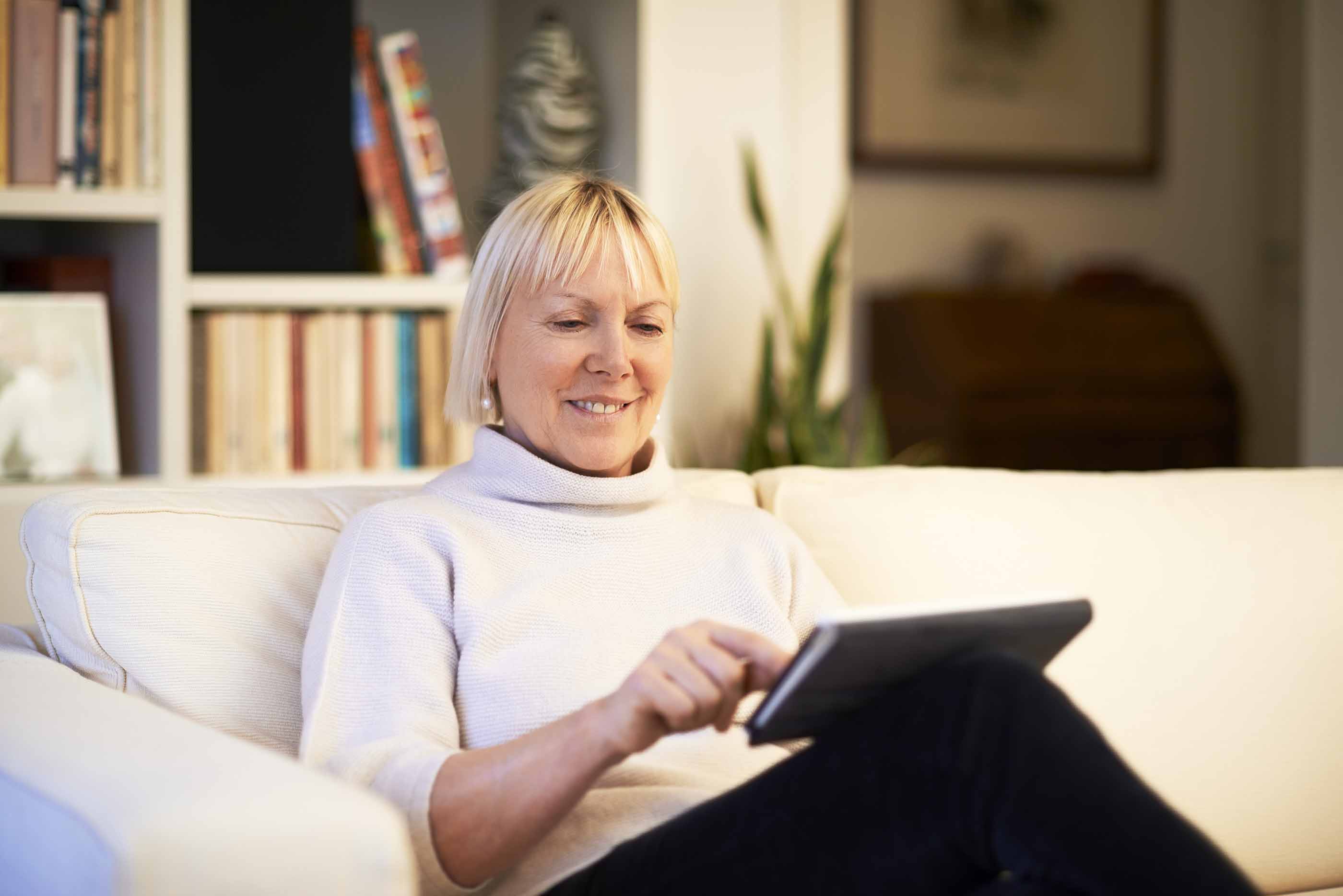 An older woman sits on a couch reading a computer tablet