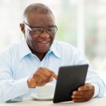 A senior Black man uses a tablet at his dining room table